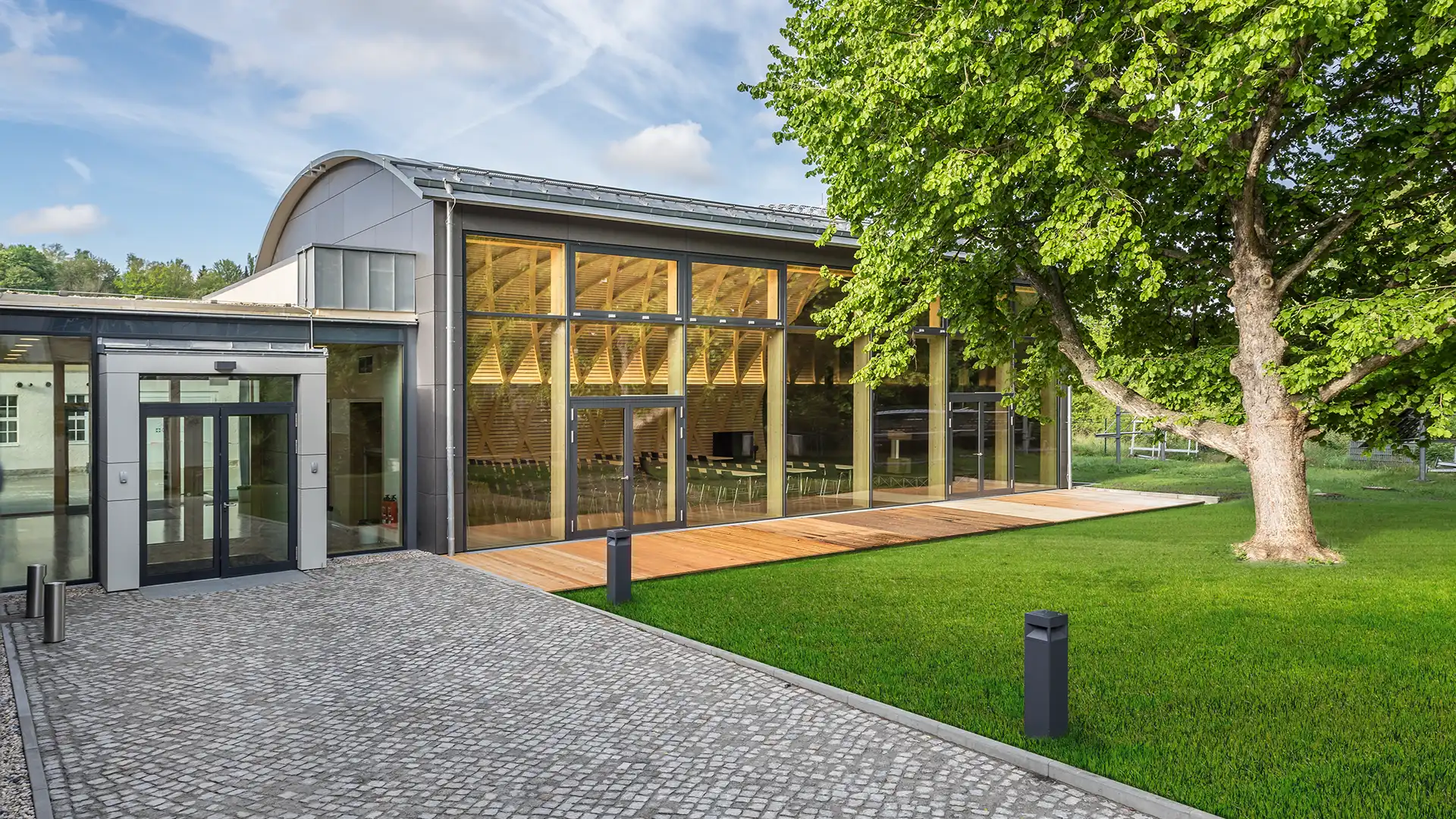 Außenansicht Konferenzzentrums mit großer Glasfront und Holzelementen. Ein markanter Haselnussbaum und eine gepflegte Rasenfläche umrahmen den Eingangsbereich und verleihen der Architektur einen natürlichen Akzent.