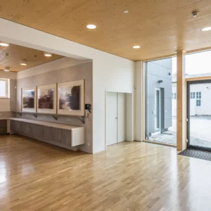 Ein großzügiges, helles Foyer mit Holzboden und Holzdecke. Große Glasflächen lassen viel Tageslicht herein, während Wandbilder und eine schlichte Einrichtung den modernen Charakter des Raums unterstreichen.