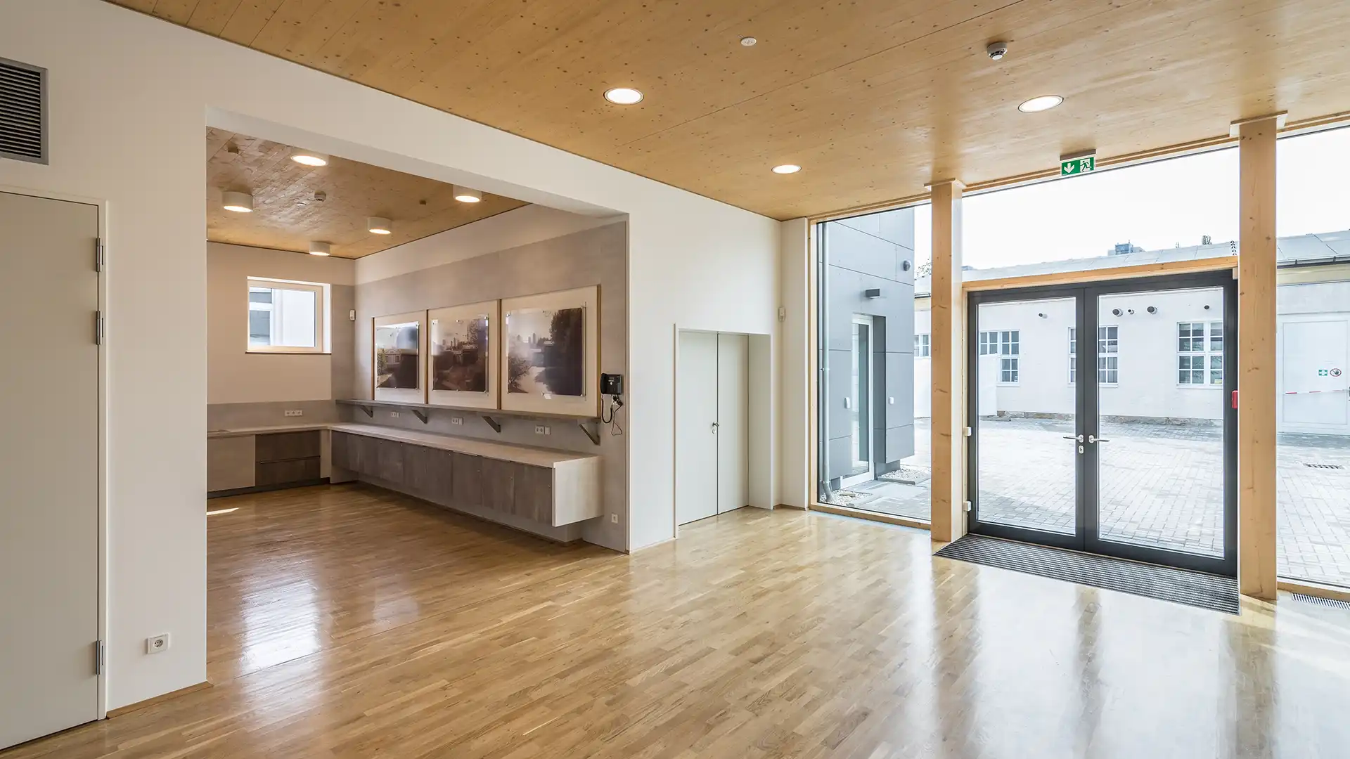 Ein großzügiges, helles Foyer mit Holzboden und Holzdecke. Große Glasflächen lassen viel Tageslicht herein, während Wandbilder und eine schlichte Einrichtung den modernen Charakter des Raums unterstreichen.