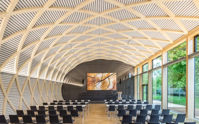 Innenansicht des modernen Konferenzraums mit beeindruckender, gewölbter Holzstruktur und großzügigen Fensterfronten. Mehrere Sitzreihen sind auf die große Monitorwand ausgerichtet, während das helle Holz und das einfallende Tageslicht eine freundliche Atmosphäre schaffen.