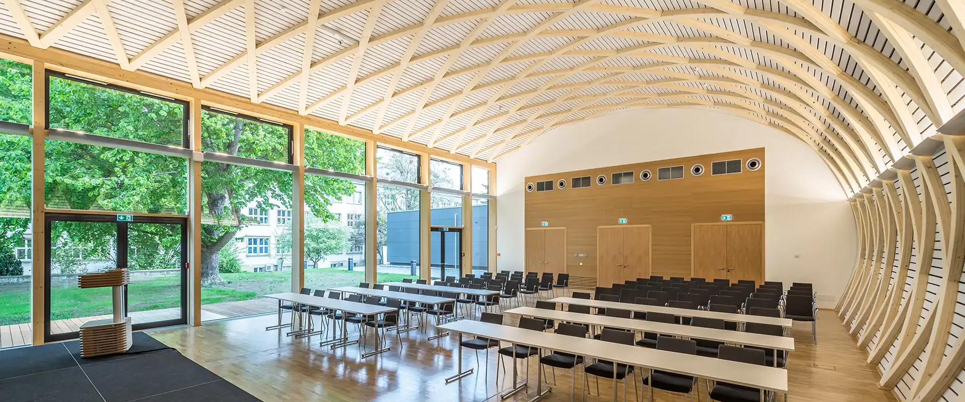Ein moderner Konferenzraum mit gebogener Holzdecke, großen Fensterfronten und variabel angeordneten Tischen und Stühlen. Die Holzstrukturen und das einfallende Tageslicht verleihen dem Raum eine helle, natürliche Atmosphäre.