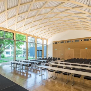 Innenansicht des Konferenzraums zur großen Fensterfront mit gebogener Holzdecke und Reihen von Tischen und Stühlen. Die Holzstrukturen und das einfallende Tageslicht verleihen dem Raum eine helle, natürliche Atmosphäre.