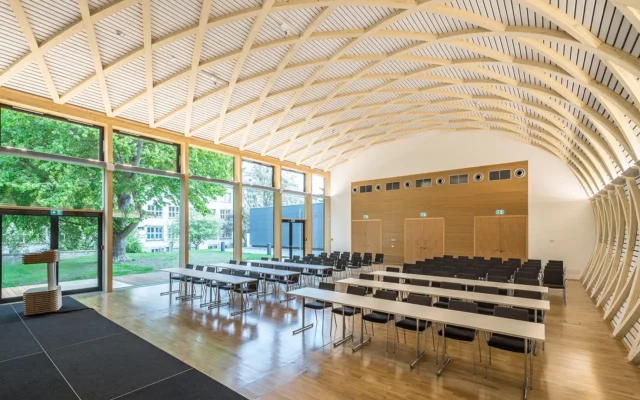 Ein moderner Konferenzraum mit gebogener Holzdecke, großen Fensterfronten und variabel angeordneten Tischen und Stühlen. Die Holzstrukturen und das einfallende Tageslicht verleihen dem Raum eine helle, natürliche Atmosphäre.