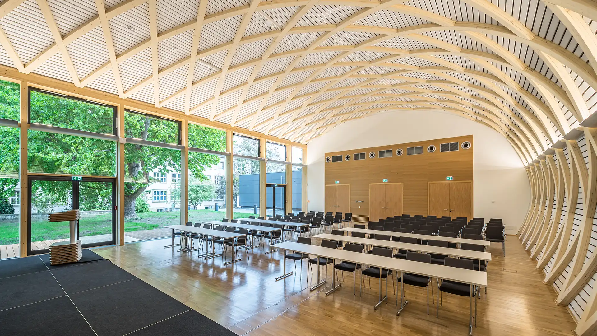 Innenansicht des Konferenzraums zur großen Fensterfront mit gebogener Holzdecke und Reihen von Tischen und Stühlen. Die Holzstrukturen und das einfallende Tageslicht verleihen dem Raum eine helle, natürliche Atmosphäre.