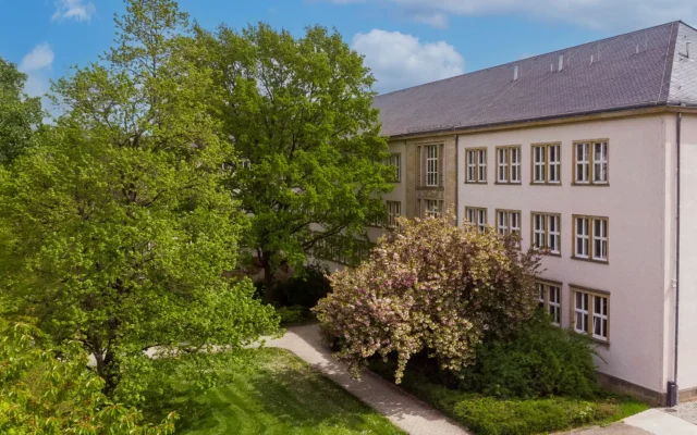 Außenansicht des IHD Dresden umgeben von Grünflächen mit blühenden Bäumen und Spazierweg.