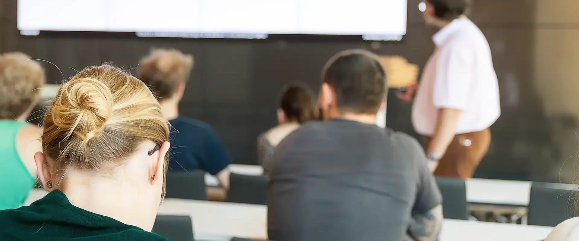 Blick von hinten auf Zuhörer in einem Seminarraum, die einem Vortragenden vor einer großen Bildschirmprojektion folgen.