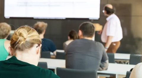 View from behind of people in a seminar room listening to a speaker in front of a large screen projection.