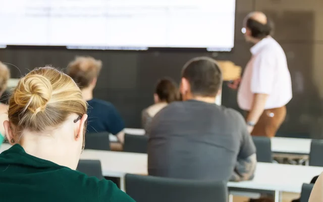 Blick von hinten auf Zuhörer in einem Seminarraum, die einem Vortragenden vor einer großen Bildschirmprojektion folgen.
