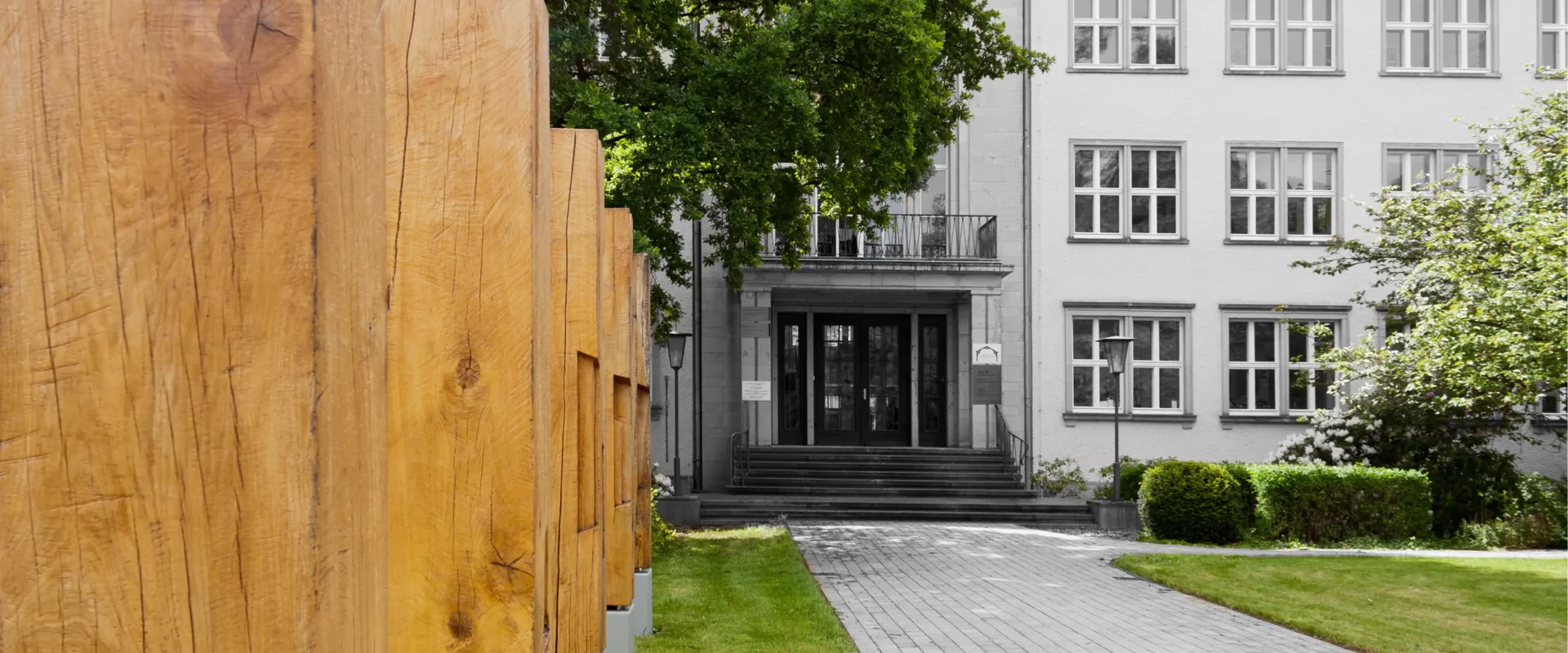 Vorgarten des IHD Dresden: Reihe massiver Eichenpfosten auf Betonfundamenten links, gepflasterter Weg führt zum Gebäudeeingang mit Treppe und mehrgeschossiger Fassade im Hintergrund.