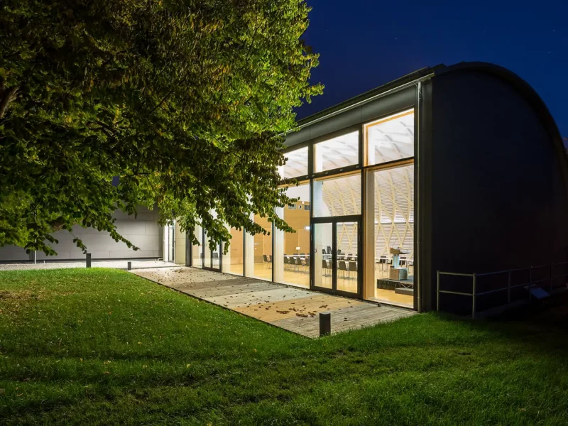 Außenansicht Konferenzzentrums bei Nacht mit großer Glasfront und Holzelementen. Ein markanter Haselnussbaum und eine gepflegte Rasenfläche umrahmen den Eingangsbereich und verleihen der Architektur einen natürlichen Akzent.