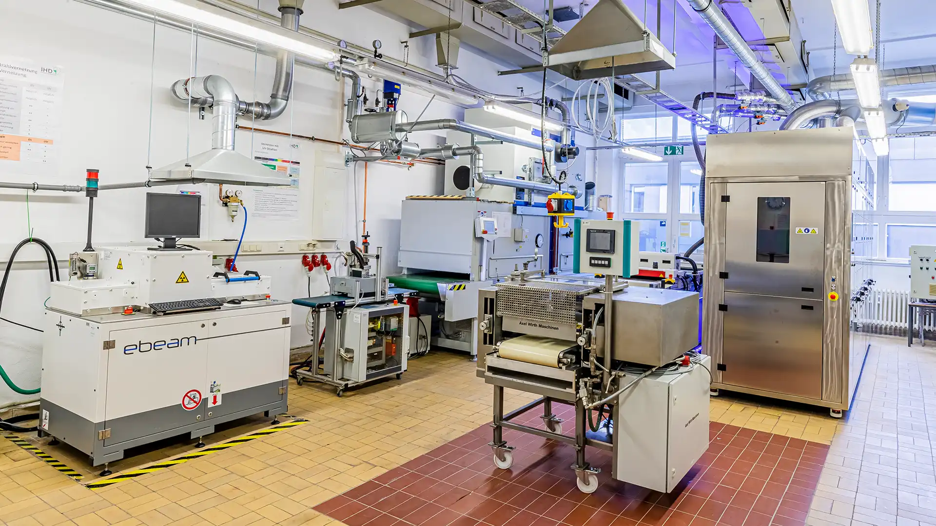 Lacklabor des IHD: Beschichtungsstrecke mit IR-Durchlaufofen, UV-Härtungsanlagen und Elektronenstrahl-(E-Beam) Anlage; Absaugungen, Steuerpulte und Fördertechnik in einem hellen Technikum.