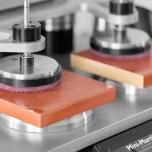 Mini-Martindale-Abriebprüfgerät in Nahaufnahme: Zwei rot lackierte Probeplatten unter rotierenden Schleifköpfen auf dem Prüfstand.