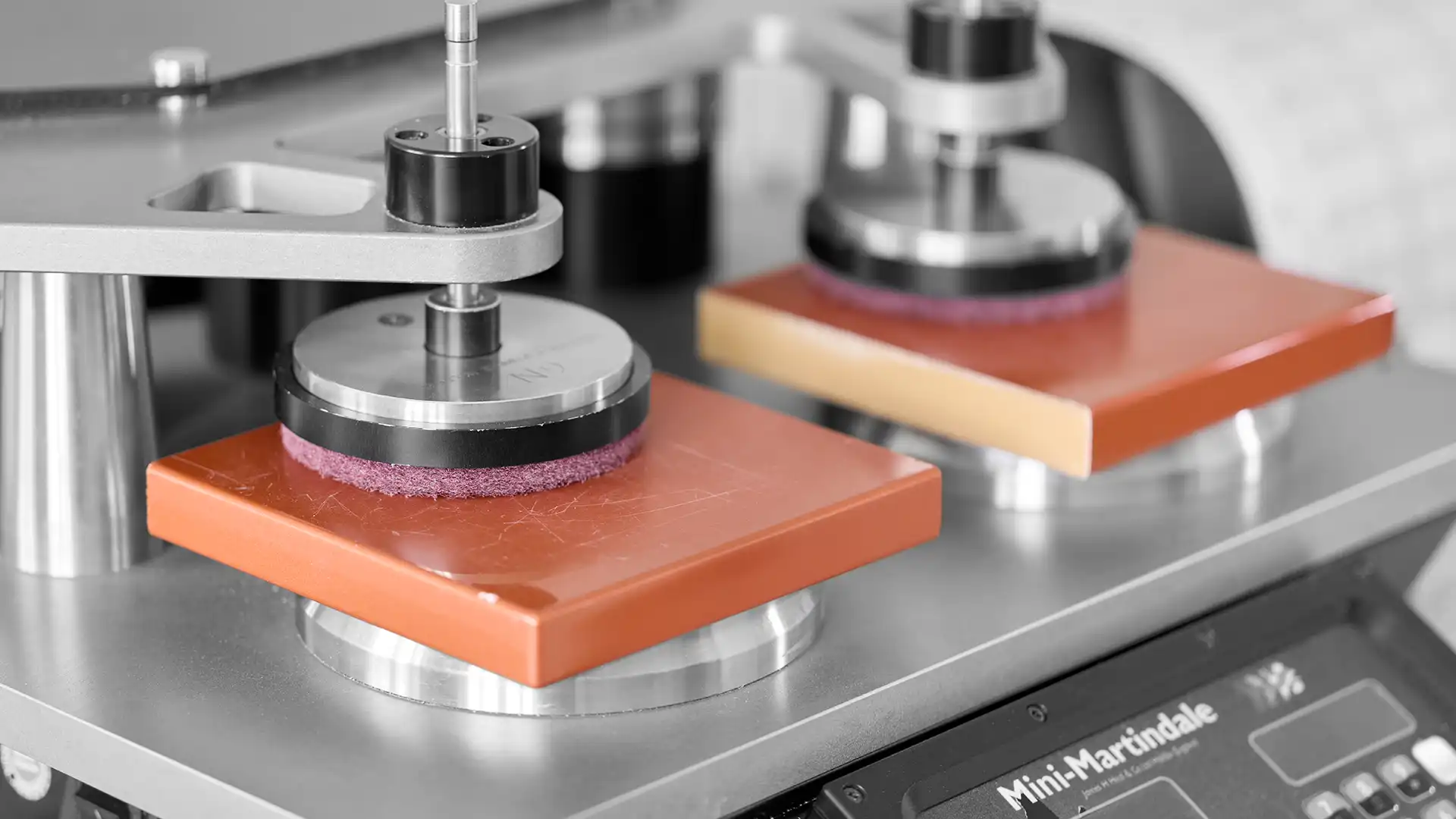 Mini-Martindale-Abriebprüfgerät in Nahaufnahme: Zwei rot lackierte Probeplatten unter rotierenden Schleifköpfen auf dem Prüfstand.