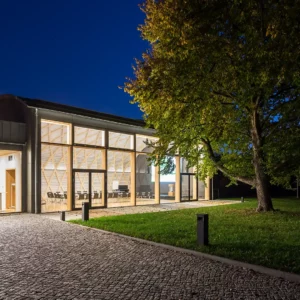 Außenansicht Konferenzzentrum bei Nacht mit großer Glasfront und Holzelementen. Ein markanter Haselnussbaum und eine gepflegte Rasenfläche umrahmen den Eingangsbereich und verleihen der Architektur einen natürlichen Akzent.