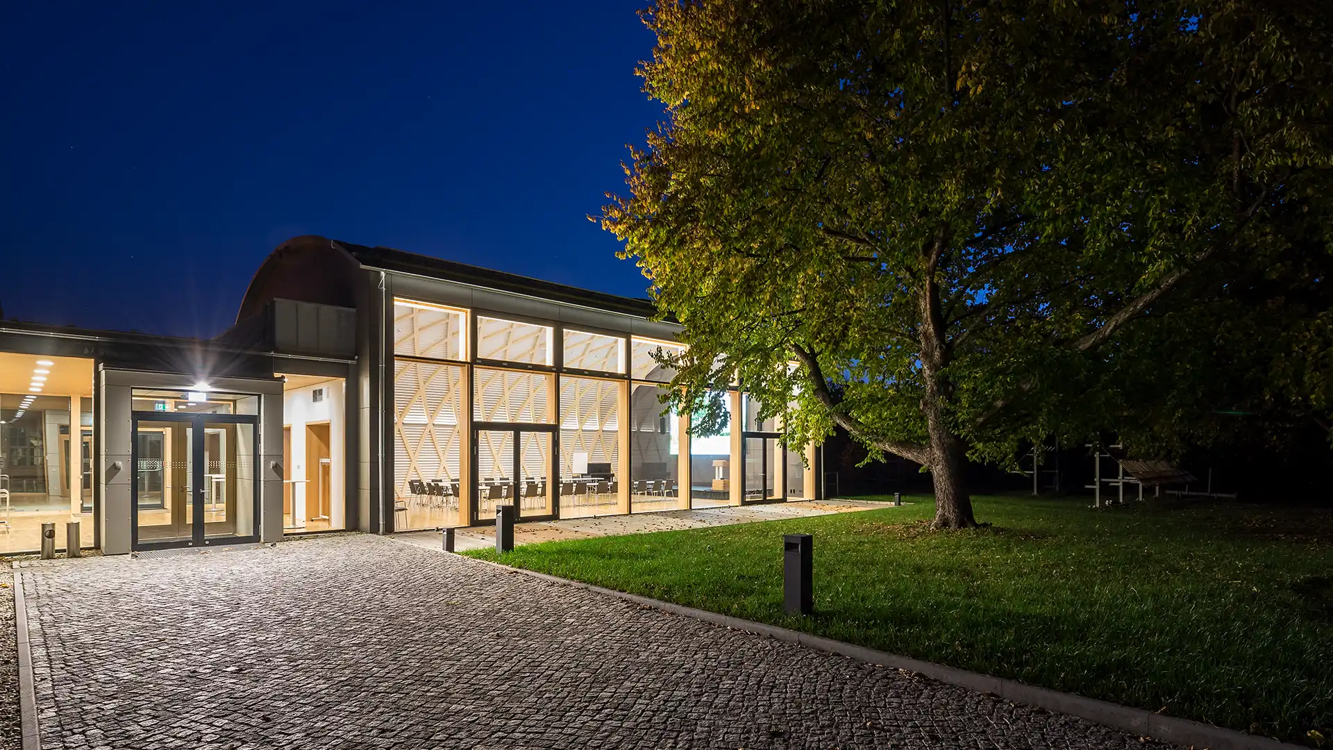 Außenansicht Konferenzzentrum bei Nacht mit großer Glasfront und Holzelementen. Ein markanter Haselnussbaum und eine gepflegte Rasenfläche umrahmen den Eingangsbereich und verleihen der Architektur einen natürlichen Akzent.