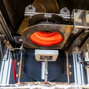 Blick in ein Cone-Kalorimeter: Unter einem glühend roten Strahlerring liegt eine Materialprobe auf der Probenträgerplattform. Umgebende Sensoren und metallische Halterungen messen verschiedene Parameter beim Brandtest.