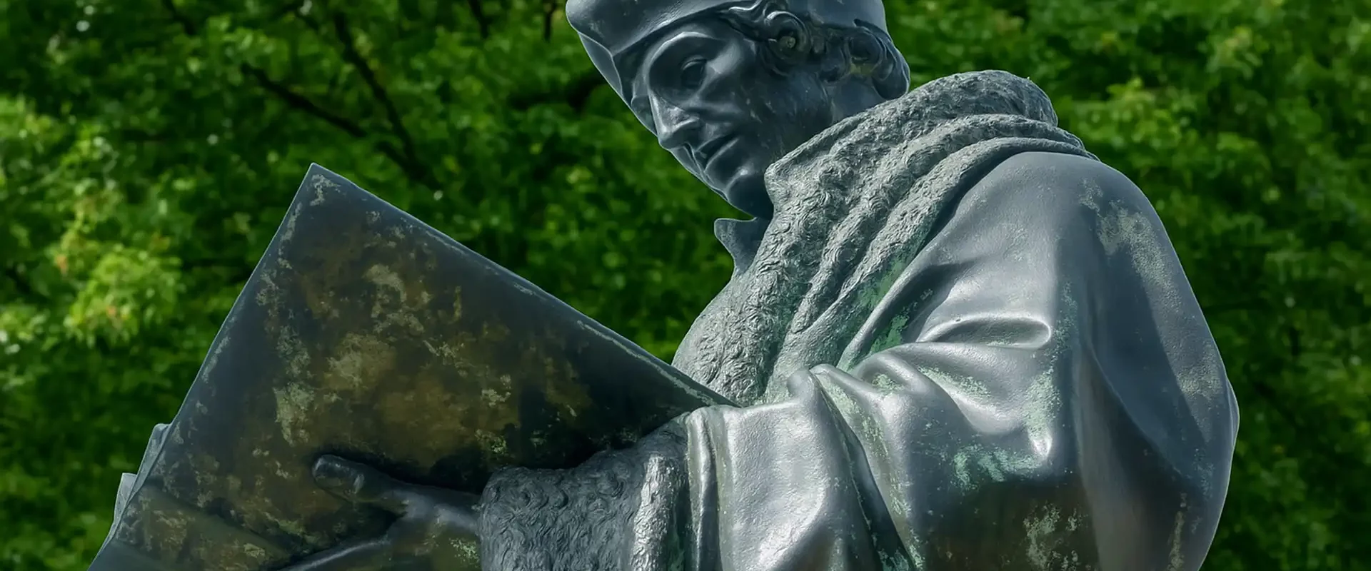 Bronzestatue des Gelehrten Erasmus von Rotterdam, den Blick auf ein aufgeschlagenes Buch gerichtet vor grün belaubtem Hintergrund.