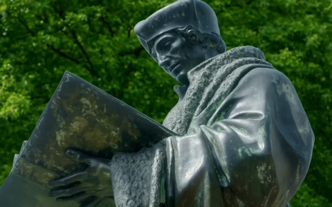 Bronzestatue des Gelehrten Erasmus von Rotterdam, den Blick auf ein aufgeschlagenes Buch gerichtet vor grün belaubtem Hintergrund.