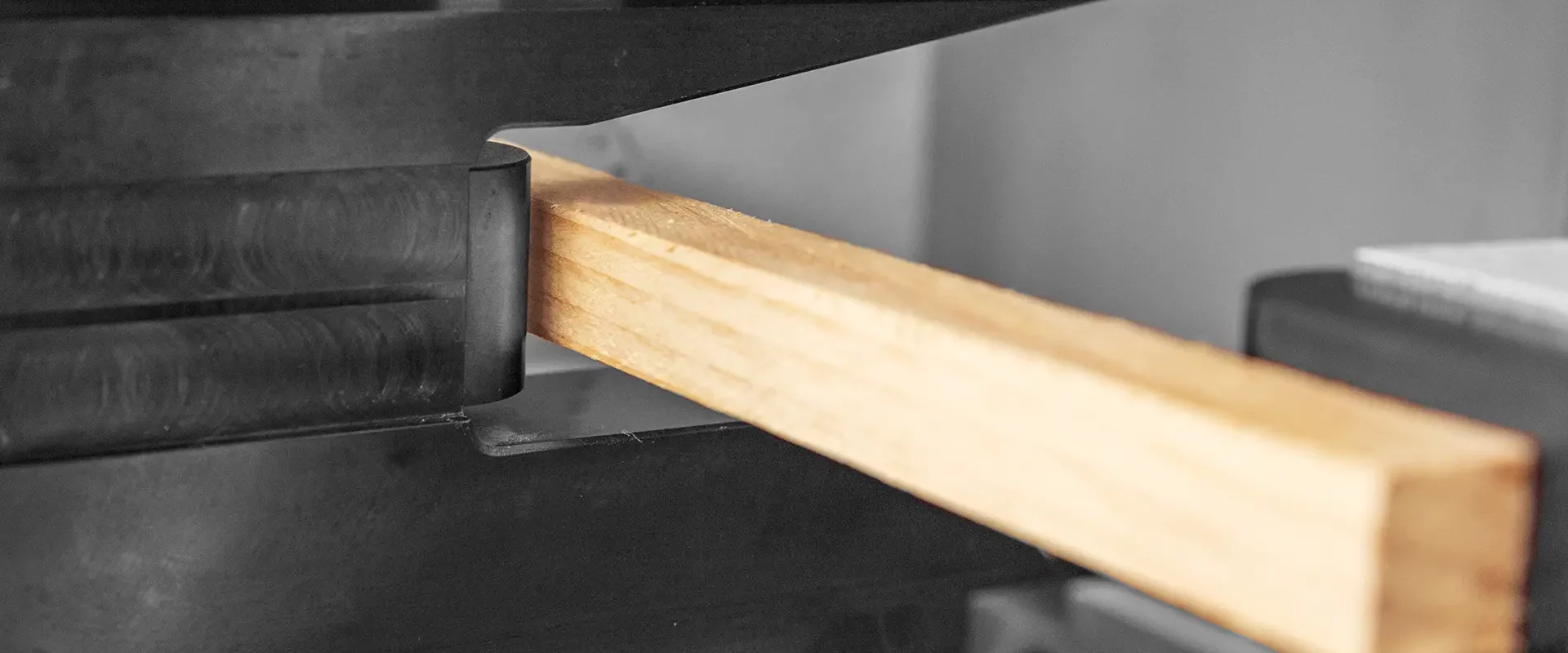 Nahaufnahme eines quadratischen Holzstabs, der in schwarzen Metallspannbacken eingespannt ist – Teil einer physikalischen Prüfaufbau zur Untersuchung mechanischer Eigenschaften.