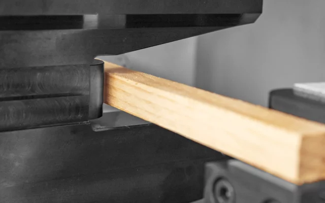 Nahaufnahme eines quadratischen Holzstabs, der in schwarzen Metallspannbacken eingespannt ist – Teil einer physikalischen Prüfaufbau zur Untersuchung mechanischer Eigenschaften.
