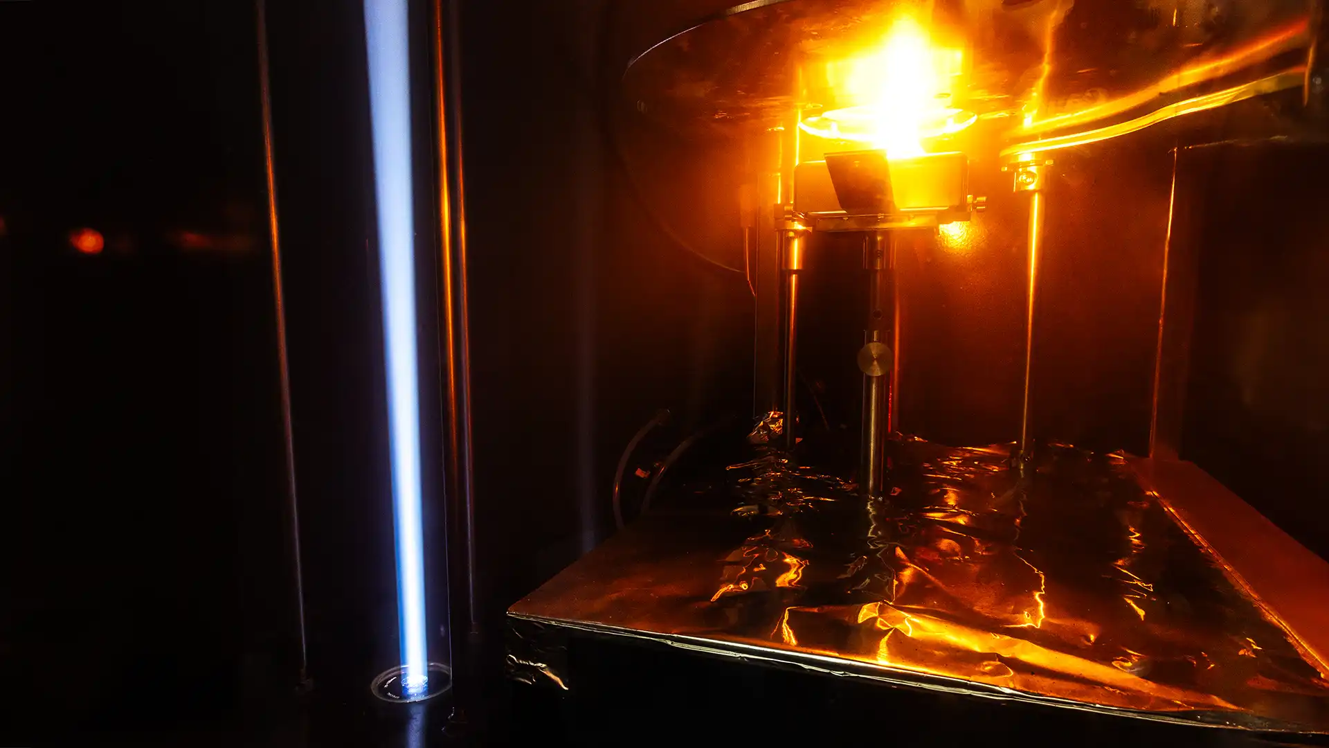 Blick in einen Rauchentwicklungsprüfstand: Eine glühende Brennerflamme erhitzt eine Materialprobe auf einer metallischen Trägerplatte, während Lichtsensoren die entstehenden Rauchgase messen.