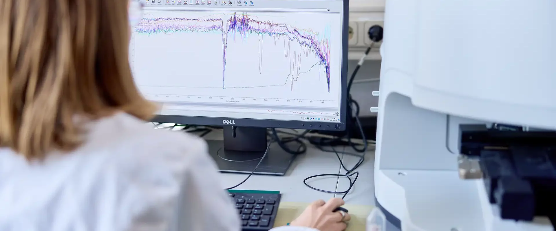 Laborantin in weißem Kittel sitzt vor einem Bildschirm, auf dem mehrfarbige Spektralkurven (FTIR-Analyse) angezeigt werden, neben einem angeschlossenen Analysegerät.