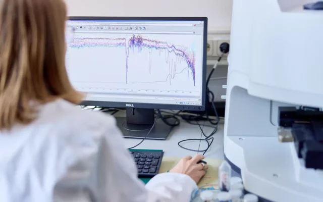 Laborantin in weißem Kittel sitzt vor einem Bildschirm, auf dem mehrfarbige Spektralkurven (FTIR-Analyse) angezeigt werden, neben einem angeschlossenen Analysegerät.