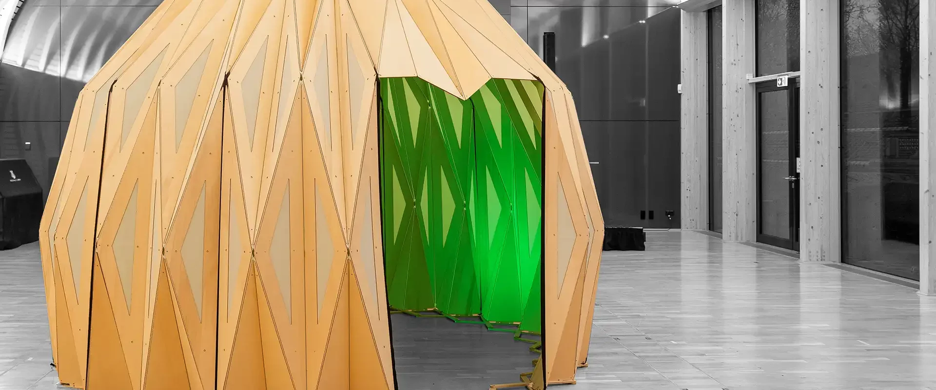 Pavillon aus gefalteten Holzpaneelen mit Eingang, innen grün beleuchtet, aufgestellt in einer Halle.