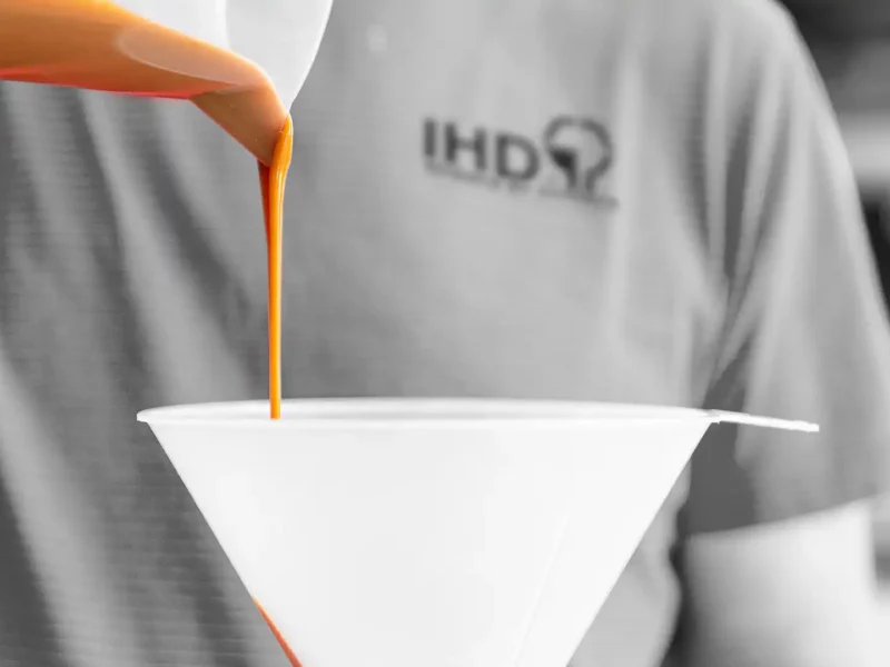 Braune Klebstoffflüssigkeit wird aus einem Becher in einen weißen Trichter gegossen, Person mit IHD Shirt im Hintergrund.