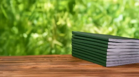 Stacked dark green brochures on a wooden surface against a blurred green natural background – symbolising sustainable publications.