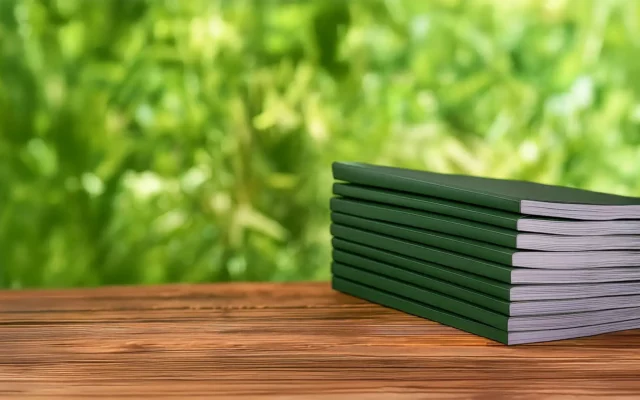 Gestapelte dunkelgrüne Broschüren auf einer Holzoberfläche vor unscharf grünem Naturhintergrund – Symbol für nachhaltige Publikationen.