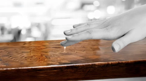 A finger touches the transparent lacquer of a richly grained wooden surface, solving small waves.