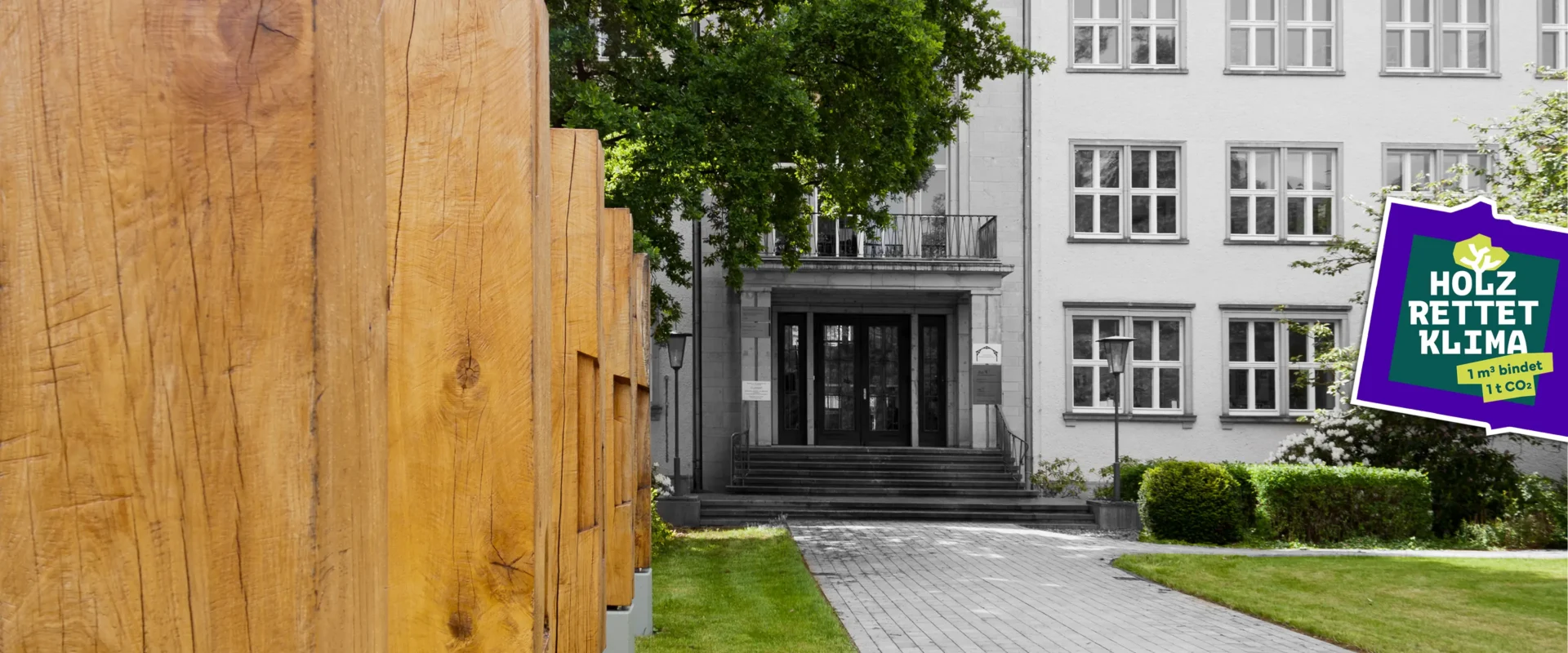 Vorgarten des IHD Dresden: Reihe massiver Eichenpfosten auf Betonfundamenten links, gepflasterter Weg führt zum Gebäudeeingang mit Treppe und mehrgeschossiger Fassade im Hintergrund. Holz Rettet Klima