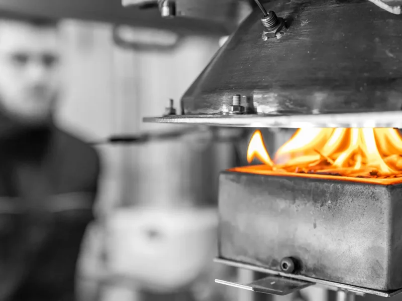 Cone-Kalorimeter im Einsatz: Eine Probe brennt mit sichtbaren Flammen im Probenhalter, darüber der konische Strahlungsheizer als Wärmeflussquelle; im Hintergrund unscharf ein Prüftechniker.